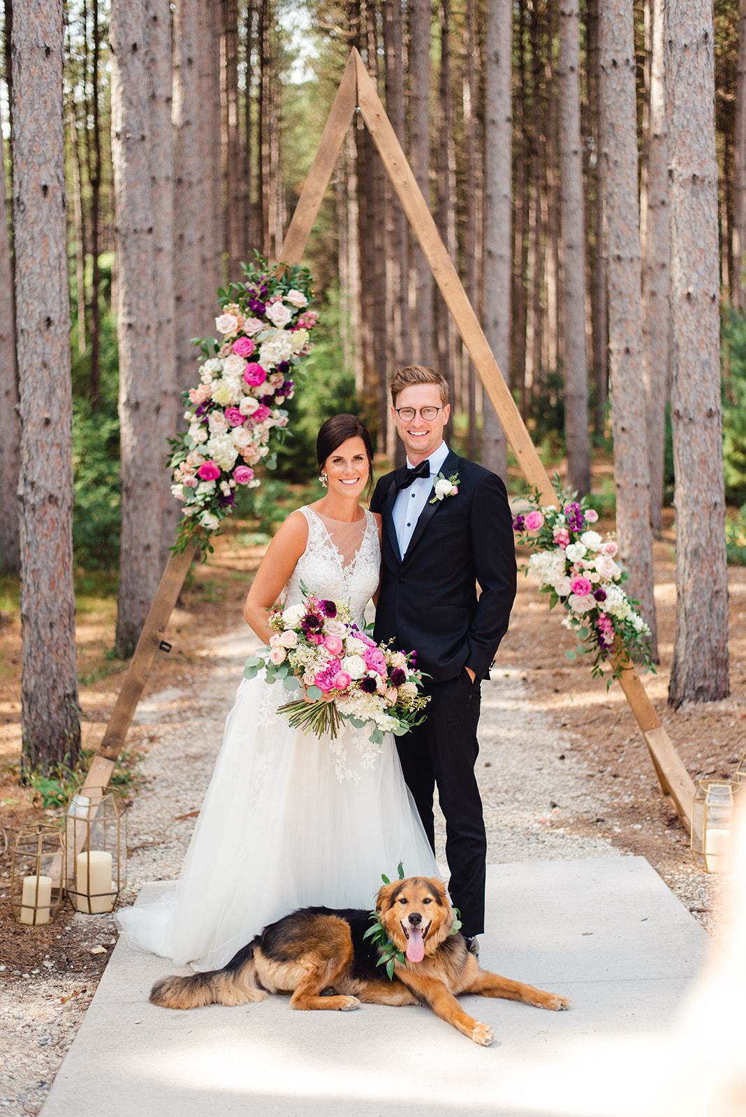 Newlyweds stand under their arbor in front of a path through pine trees with their dog laying at their feet