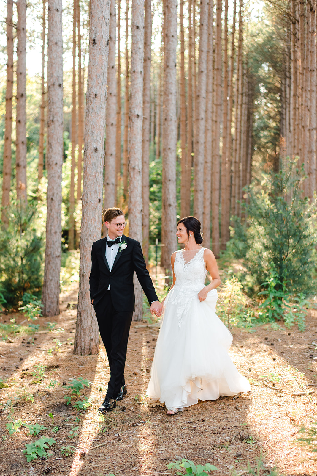 Newlyweds walk through a pine tree farm holding hands in a lace dress and black suit at a Unique Wedding Venues Minnesota