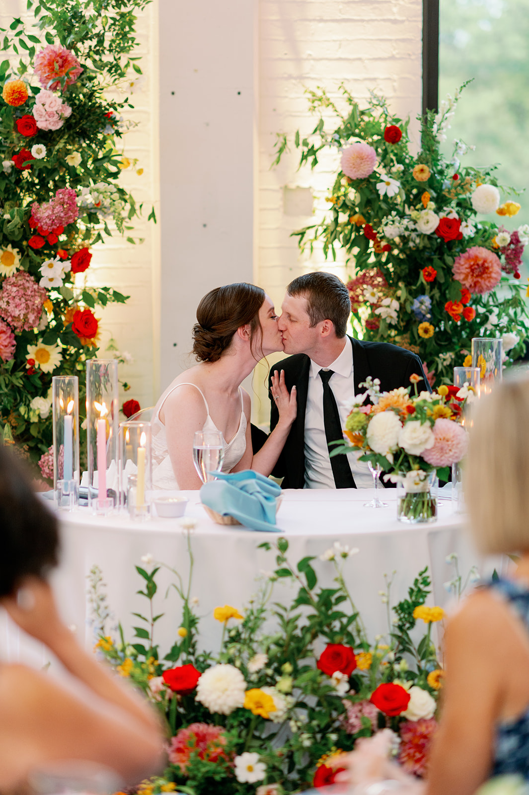 Newlyweds kiss while sitting at their reception table surrounded by colorful flowers