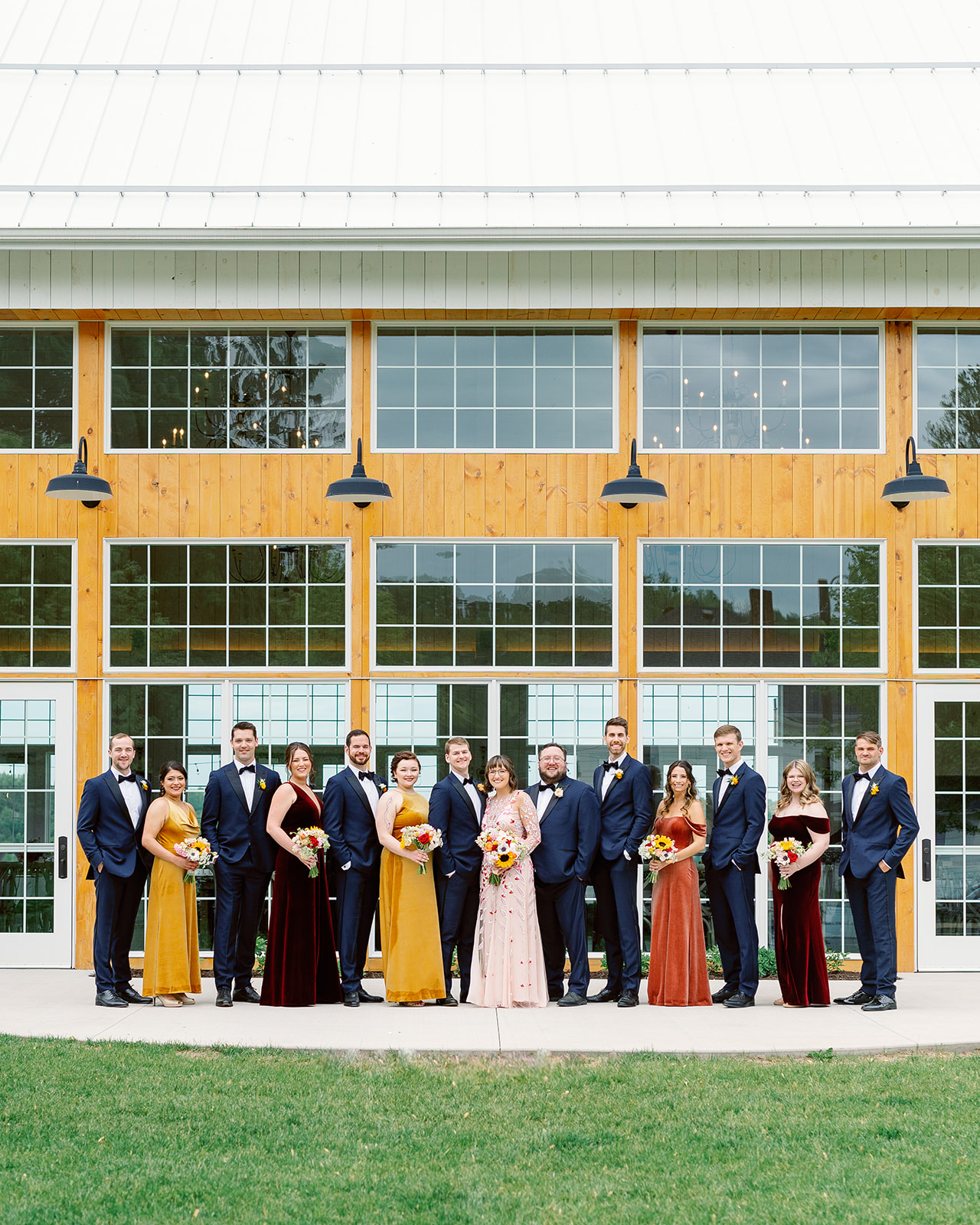 A large wedding party stands around the newlyweds outside a large Unique Wedding Venues Minnesota
