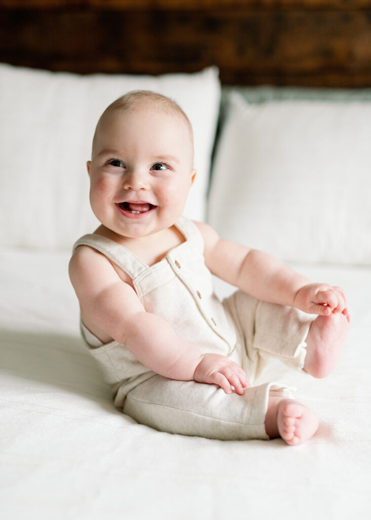 6 month old baby boy sitting on bed, grabbing his toes and smiling