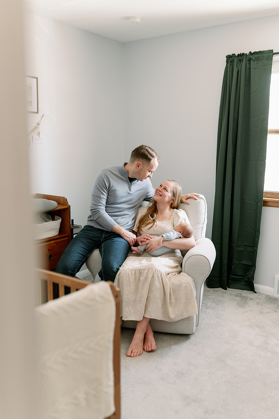Mom sits in chair cradling newborn while dad sits on the arm of the chair looking lovingly at the mom during Twin Cities newborn photography session and the best time to take newborn photos.