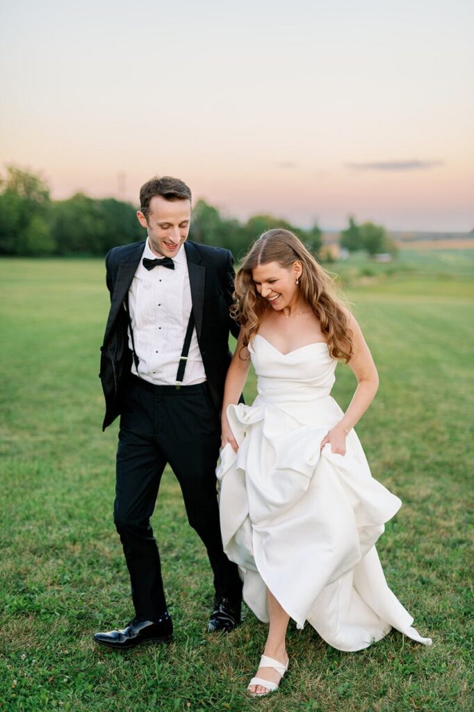 Wedding photographers in MN capture a couple walking together across a field at sunset on their wedding day. 