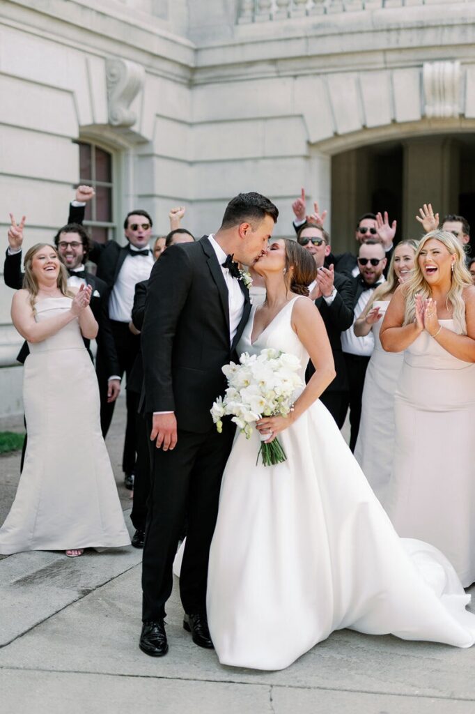 A photographer for a wedding in MN capturing a newlywed couple kissing while their wedding party celebrates around them. 