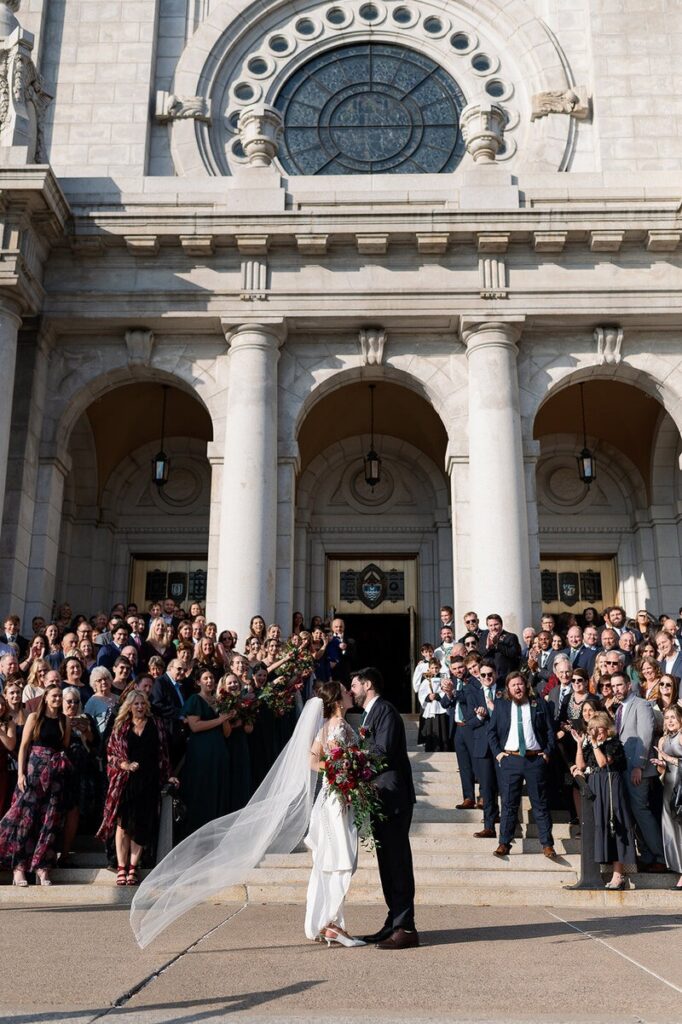 A large wedding party gathered outside a church as the couple shares a kiss, as captured by wedding photographers in MN. 